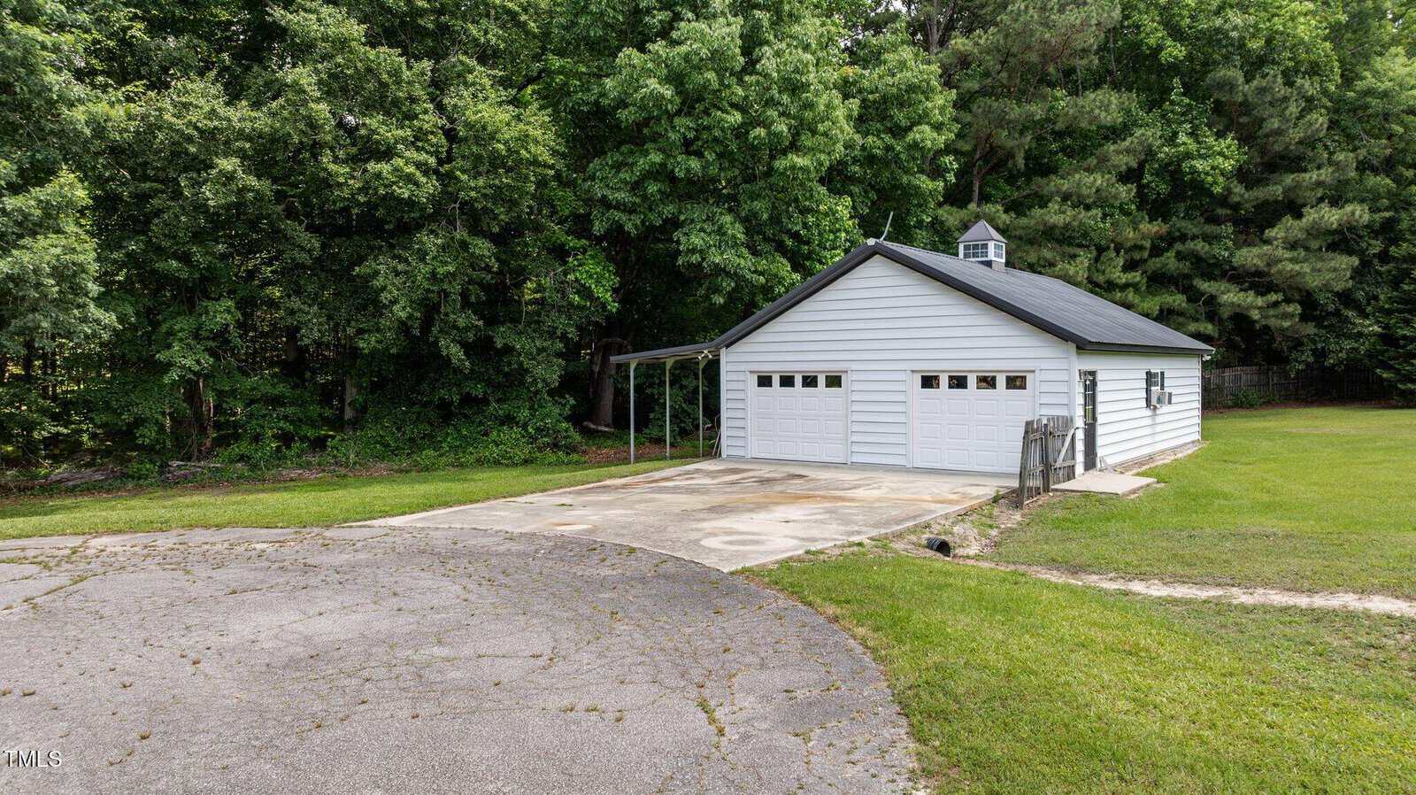 A detached white garage with three doors, a gray roof, and a small cupola sits beside a concrete driveway, surrounded by green grass and dense trees in the background.