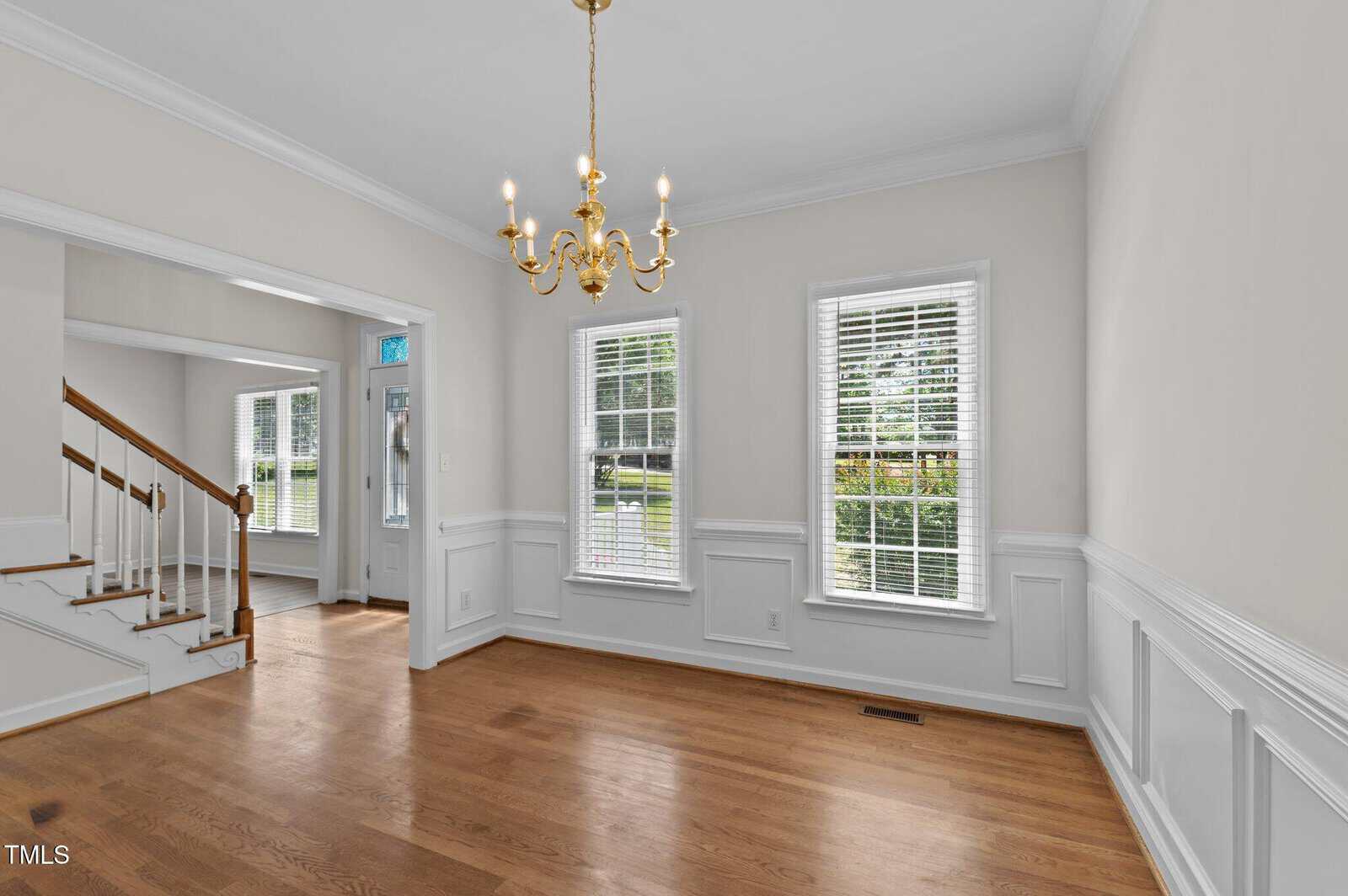 Empty room with light hardwood floors, white walls with wainscoting, three windows with blinds, a brass chandelier, and a staircase visible to the left. Natural light enters from the windows and adjacent entryway.