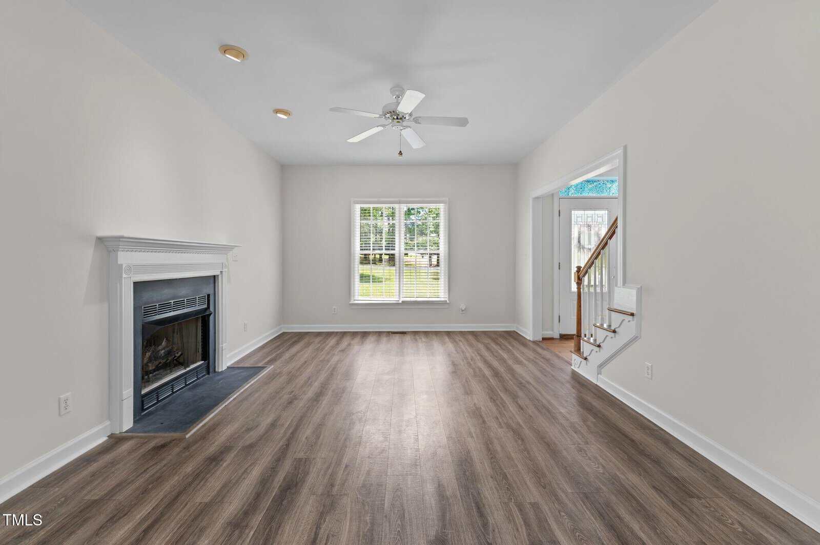 A spacious living room with light walls, wood-look flooring, a white ceiling fan, a fireplace on the left, two windows at the back, and a staircase leading up on the right.