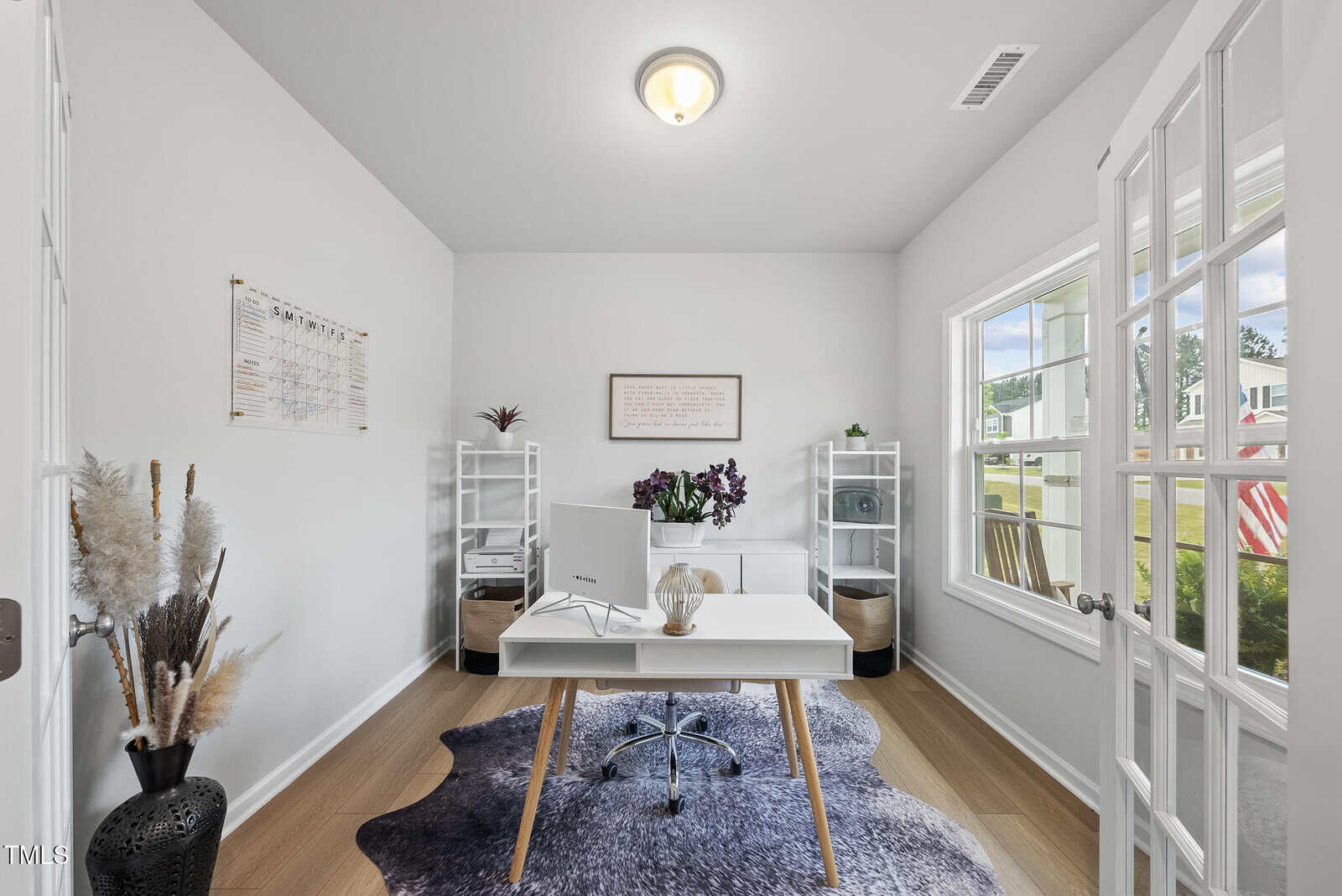 A bright home office with a desk, computer, and chair in the center, flanked by white shelves, a large window on the right, decorative plants, and wall art. The room features light walls and wood flooring.
