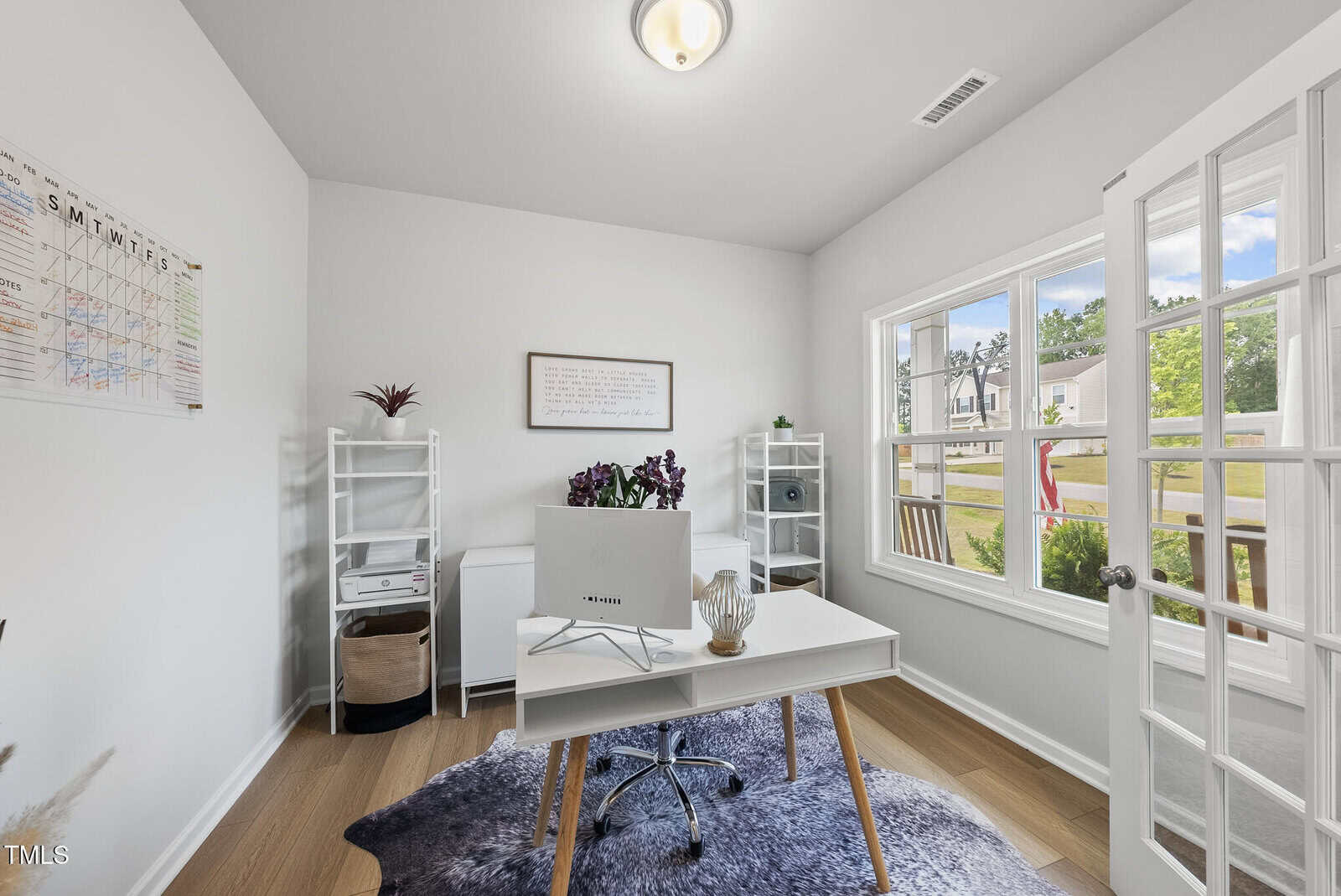 A bright home office with a white desk, computer monitor, two white shelves, a wall calendar, and a window showing a view of a porch and neighborhood. A rug and small plants are present.