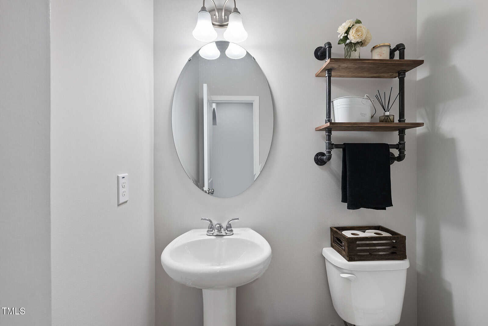 A small bathroom with a pedestal sink, round mirror, toilet, and two wooden shelves holding decor items and towels. The walls are light gray, and there is a light fixture above the mirror.