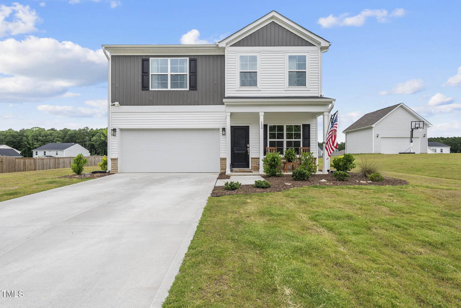 Two-story suburban house with a two-car garage, front porch, American flag, and neatly landscaped yard. The home features gray and white siding and is situated on a spacious green lawn under a partly cloudy sky.