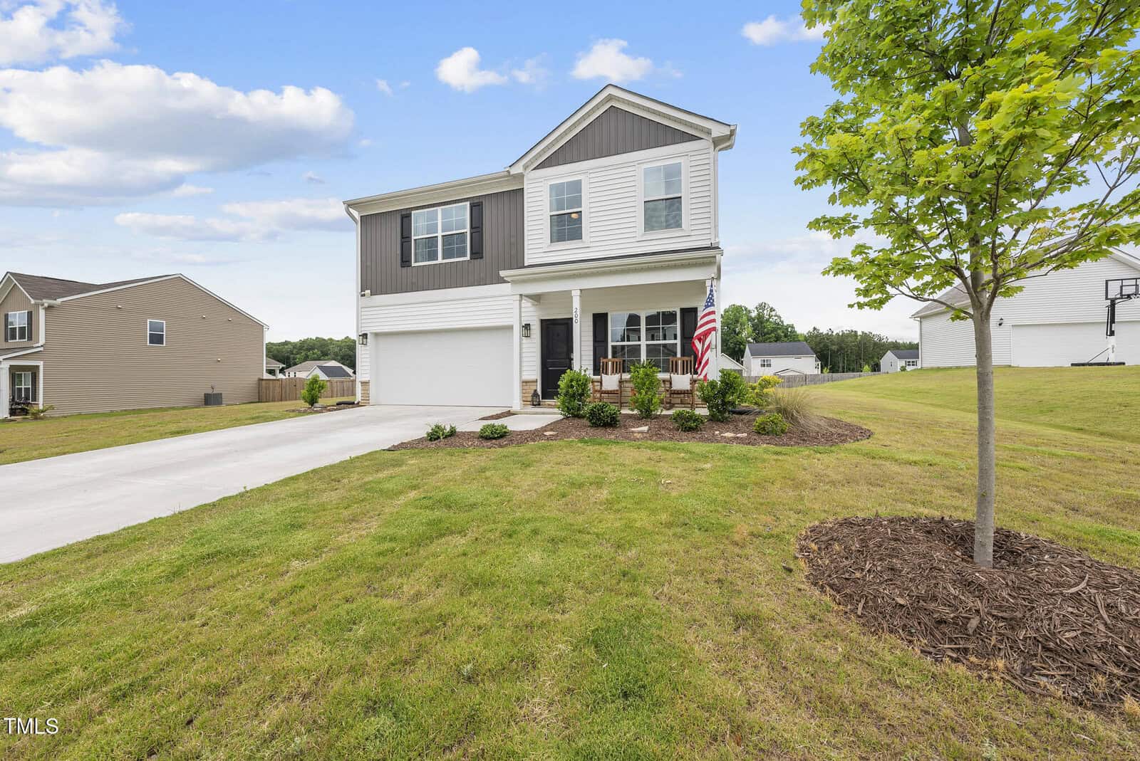 Two-story suburban house with a gray and white exterior, attached garage, U.S. flag by the front door, and a well-maintained lawn with a young tree in the yard under a partly cloudy sky.