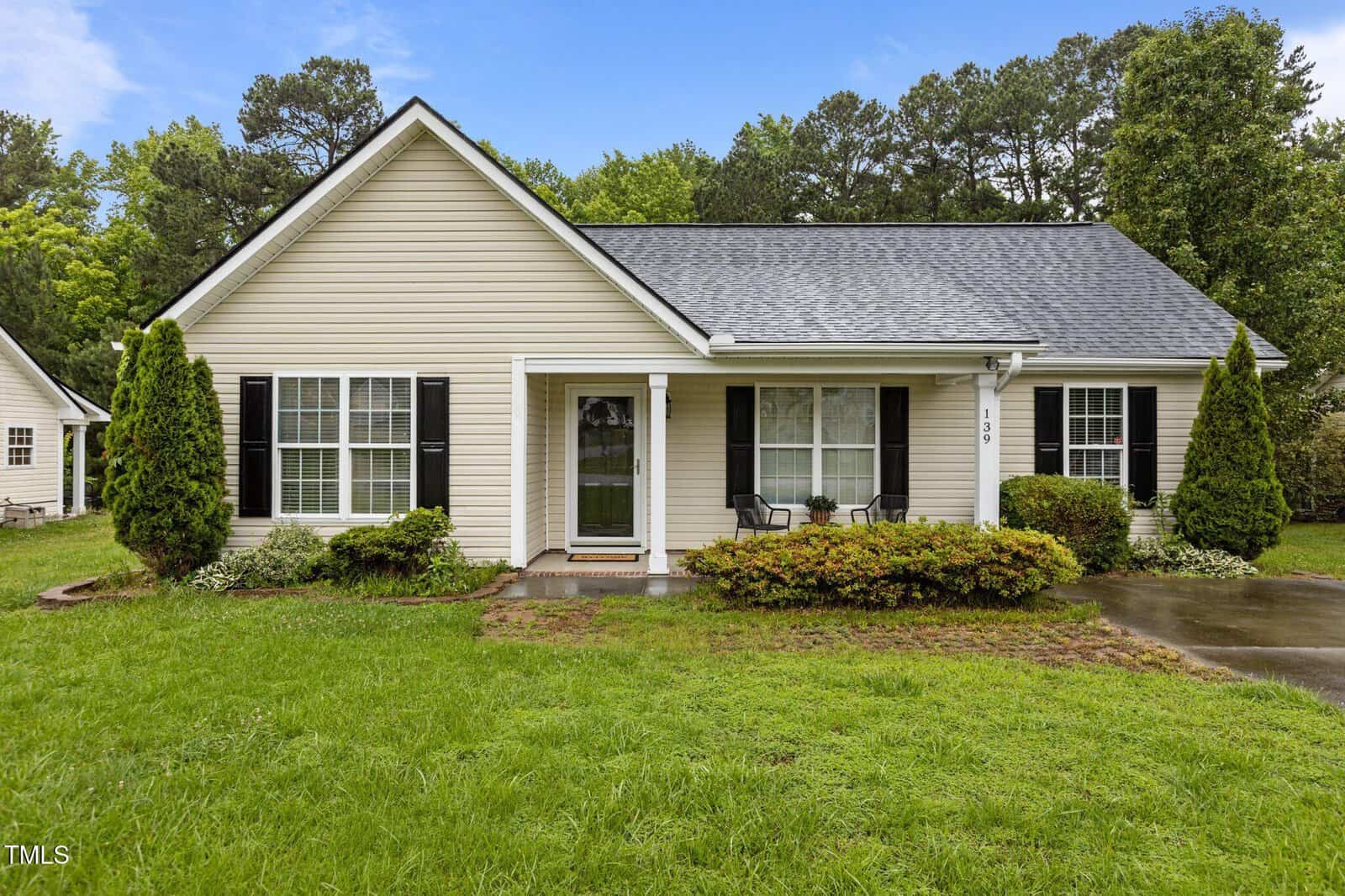 A single-story beige house with black shutters sits at 139 Carriage Hill Drive. A gable roof and small covered entry shelter two chairs on the front porch, surrounded by bushes, a grassy yard, and trees under a blue Stem sky.