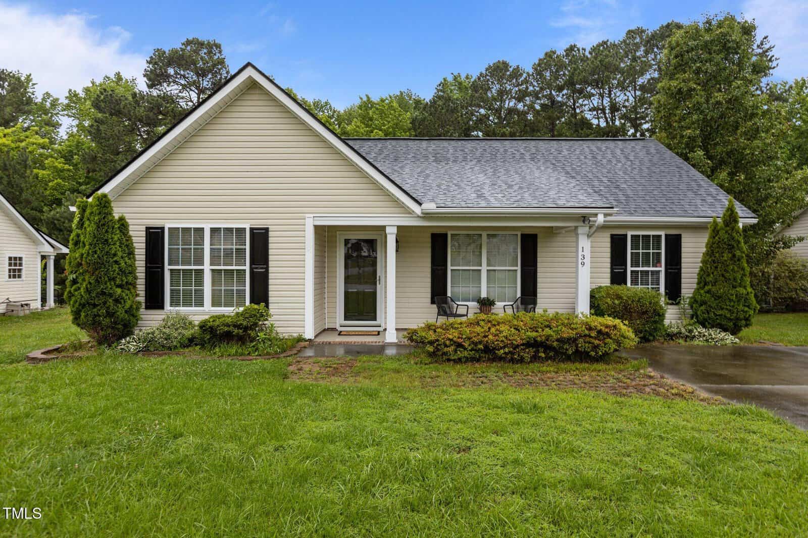 Single-story beige house with black shutters, white trim, and a gray shingle roof, featuring a small covered front porch with two chairs, shrubs, and a grassy yard bordered by trees in the background.
