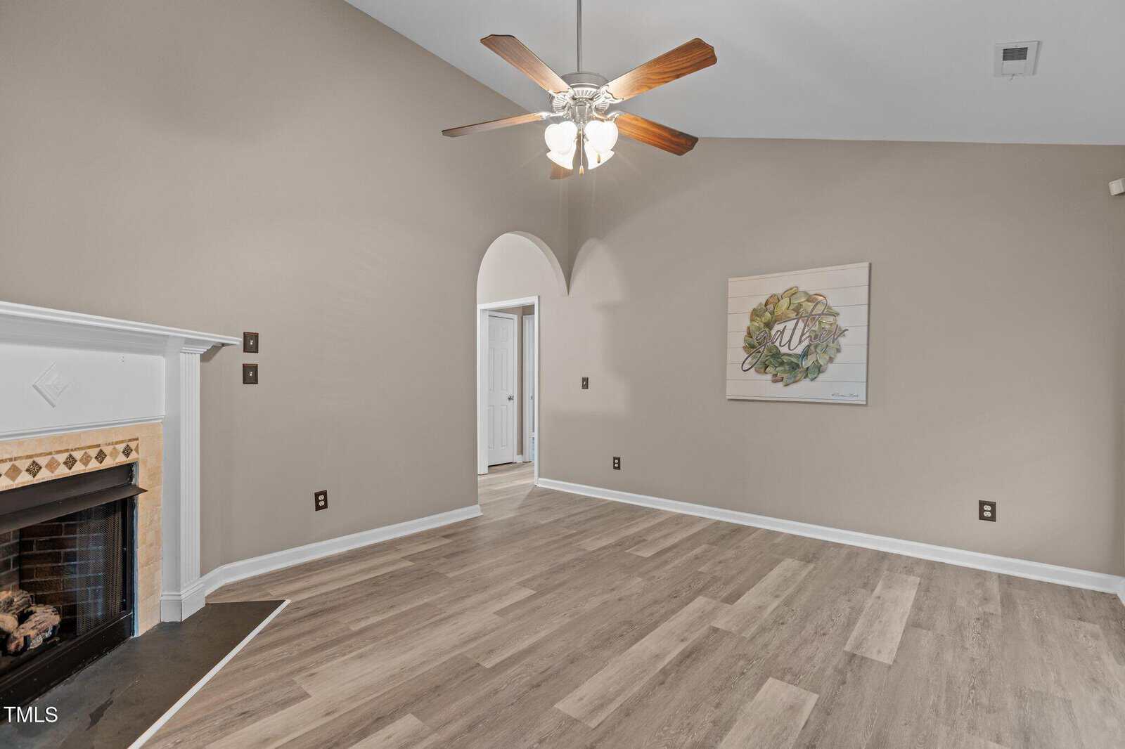 A room with light wood flooring, beige walls, a white ceiling fan with lights, a fireplace on the left, an arched doorway, and a decorative wall art piece on the right wall.