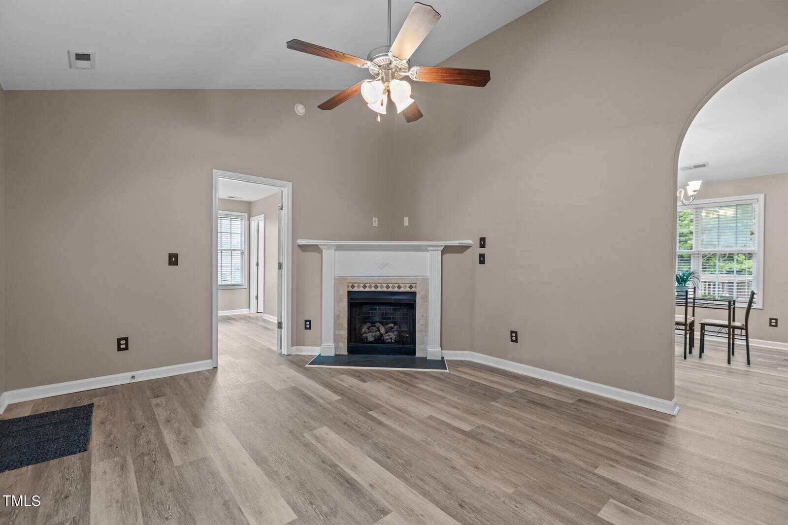 A living room with light wood flooring, beige walls, a white fireplace, and a ceiling fan with lights. There is an arched doorway leading to a dining area with windows in the background.