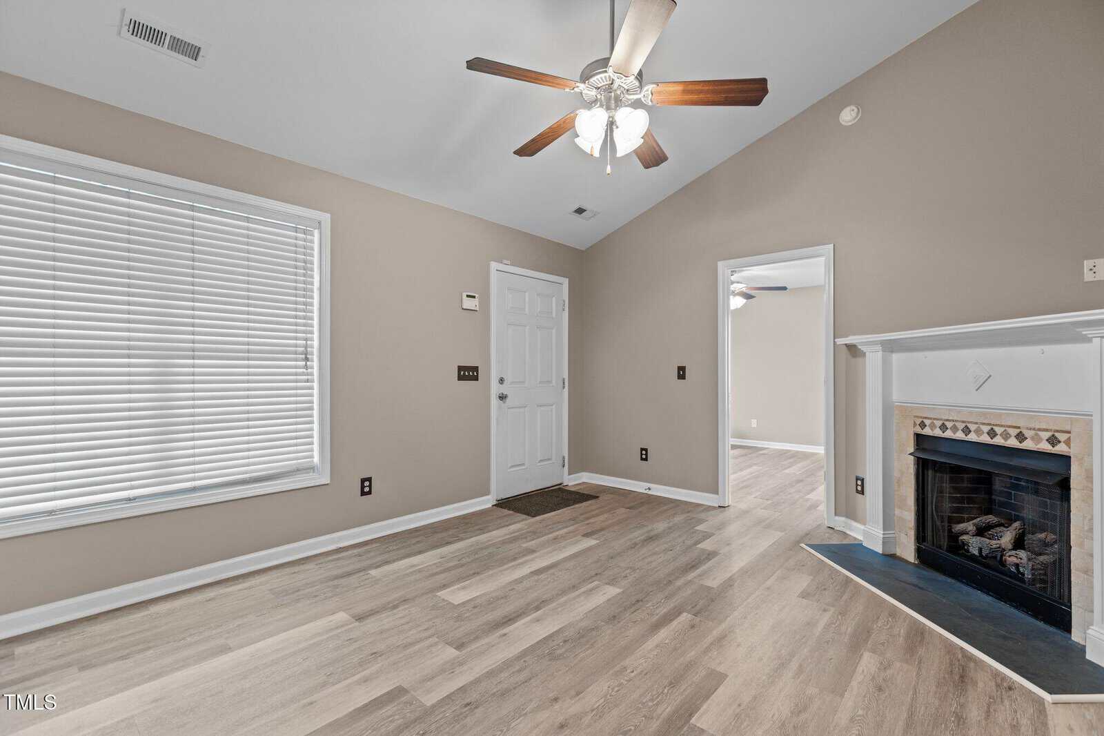A room with light wood flooring, beige walls, a ceiling fan with lights, a large window with blinds, a white front door, and a white fireplace with a black hearth. An open doorway leads to another room.