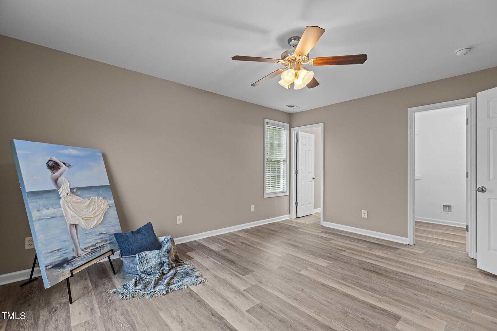 A clean, empty room with tan walls, wood floor, ceiling fan with lights, two windows, two open white doors, and a painting of a woman in a white dress on an easel beside a blue blanket and pillow.