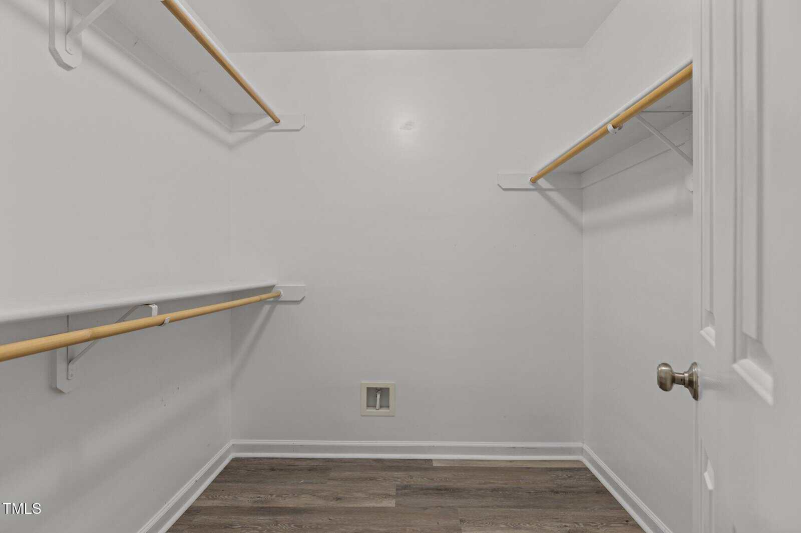 Empty walk-in closet with white walls, wooden rods, and shelves on both sides. The floor is wood-look laminate, and there is a small square wall vent near the floor at the back. The door is partially open.