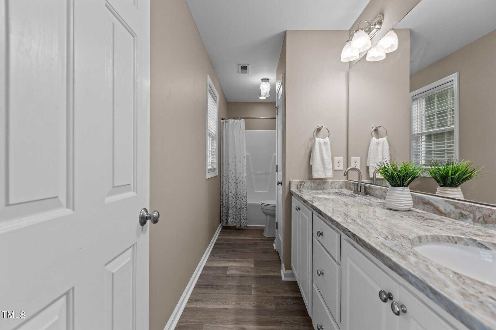 A modern bathroom with light brown walls, a double-sink marble countertop, white cabinets, wood floors, two mirrors, two small potted plants, towel rings, and a shower area with a white curtain in the back.