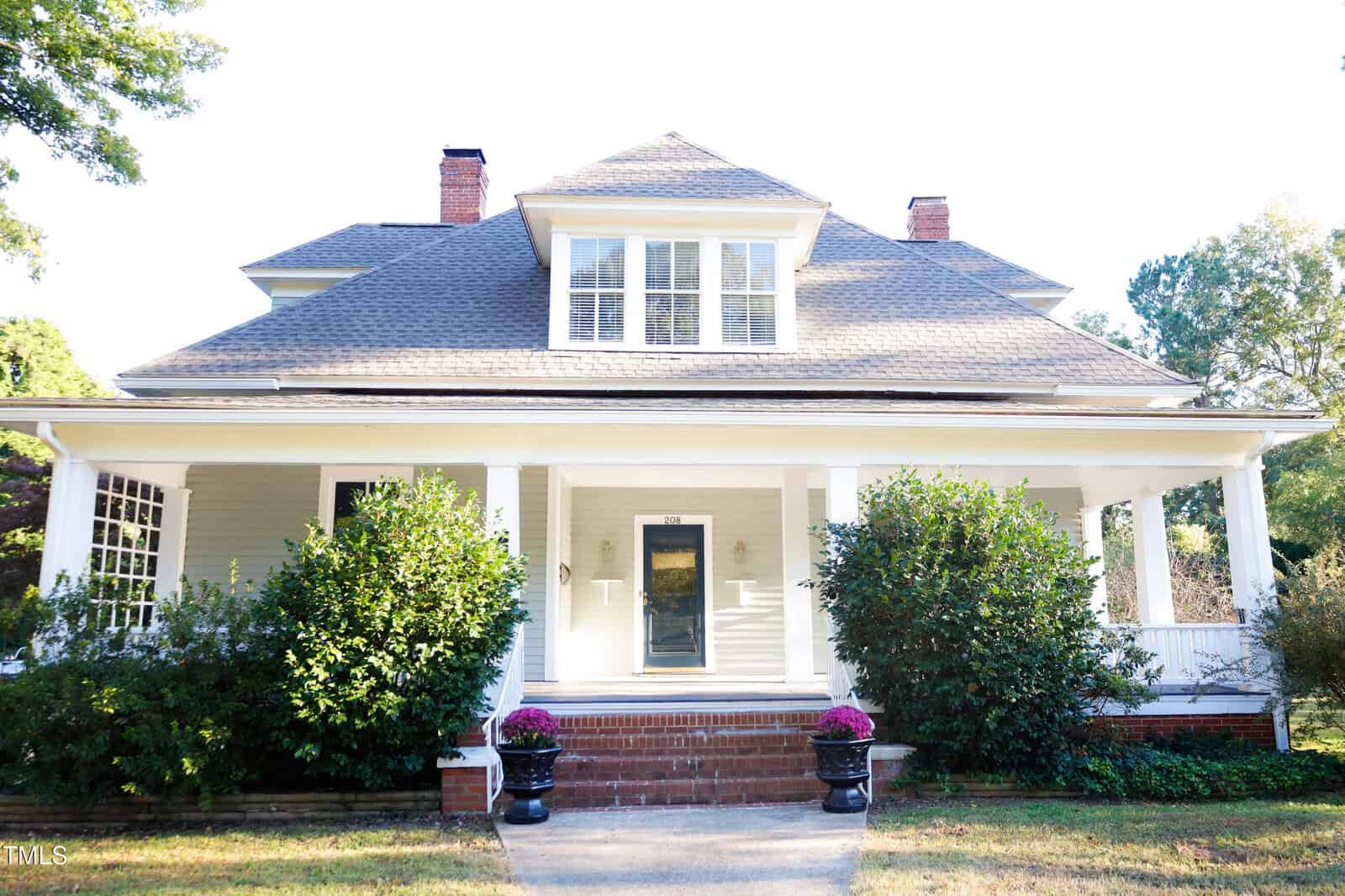 A white, two-story house with a large front porch, two brick chimneys, and a gray roof. Bushes and potted flowers line the brick steps leading to the front door. Trees surround the house, and the yard is neatly maintained.