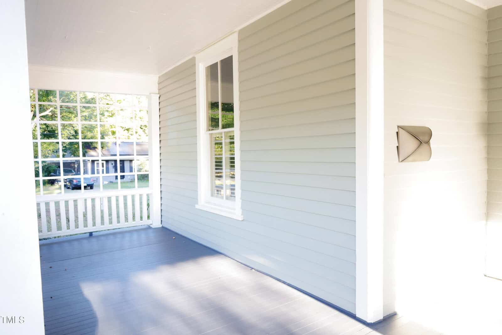 A bright, empty front porch with light gray siding, a large window, white trim, and a dark gray wooden floor. Sunlight streams in from the left, highlighting a white railing and a screen wall.