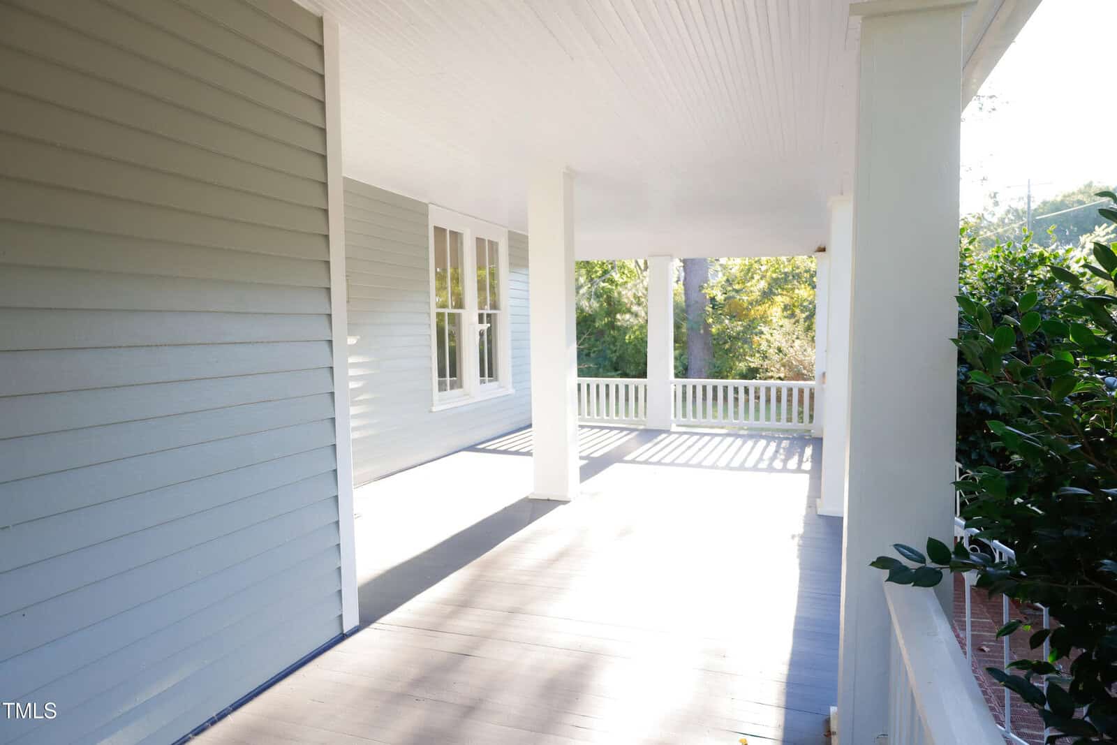 A spacious, sunlit front porch with light gray siding, white railings, and trim. Trees and greenery are visible in the background. The porch has a wooden floor and white ceiling.