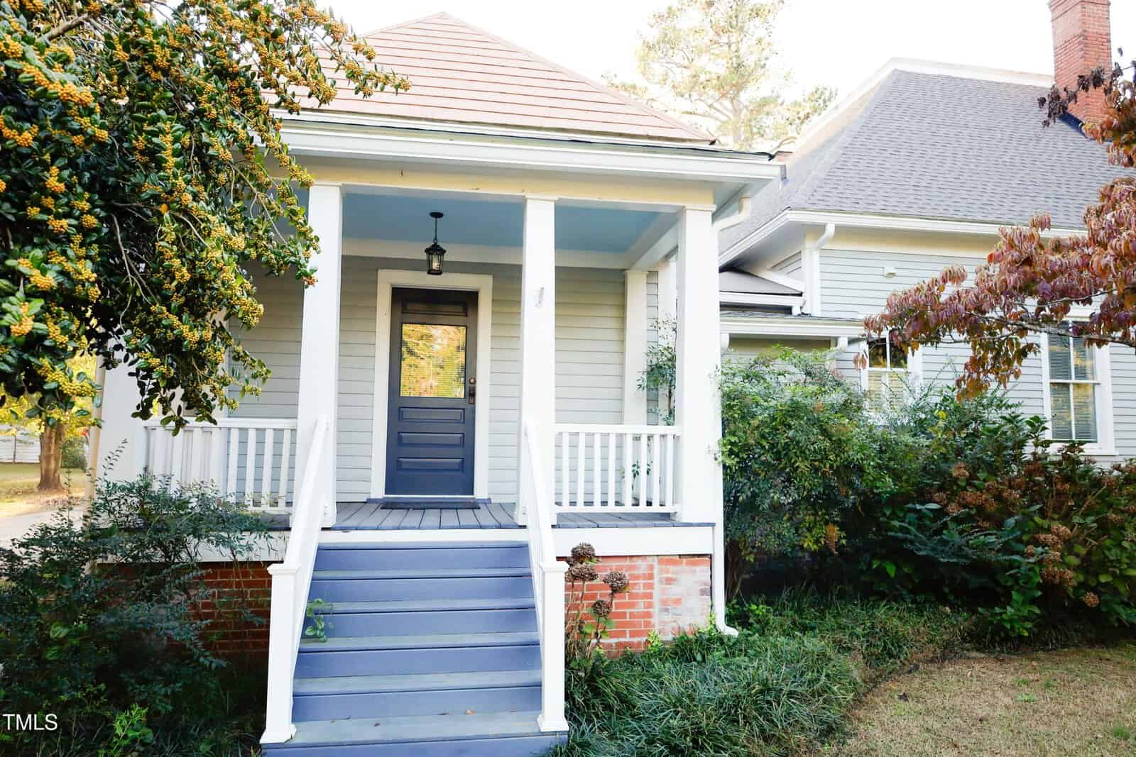 A light gray house with a small covered porch, blue steps, white columns, and trim. The yard has green shrubs and trees, and the porch features a hanging light above the front door.