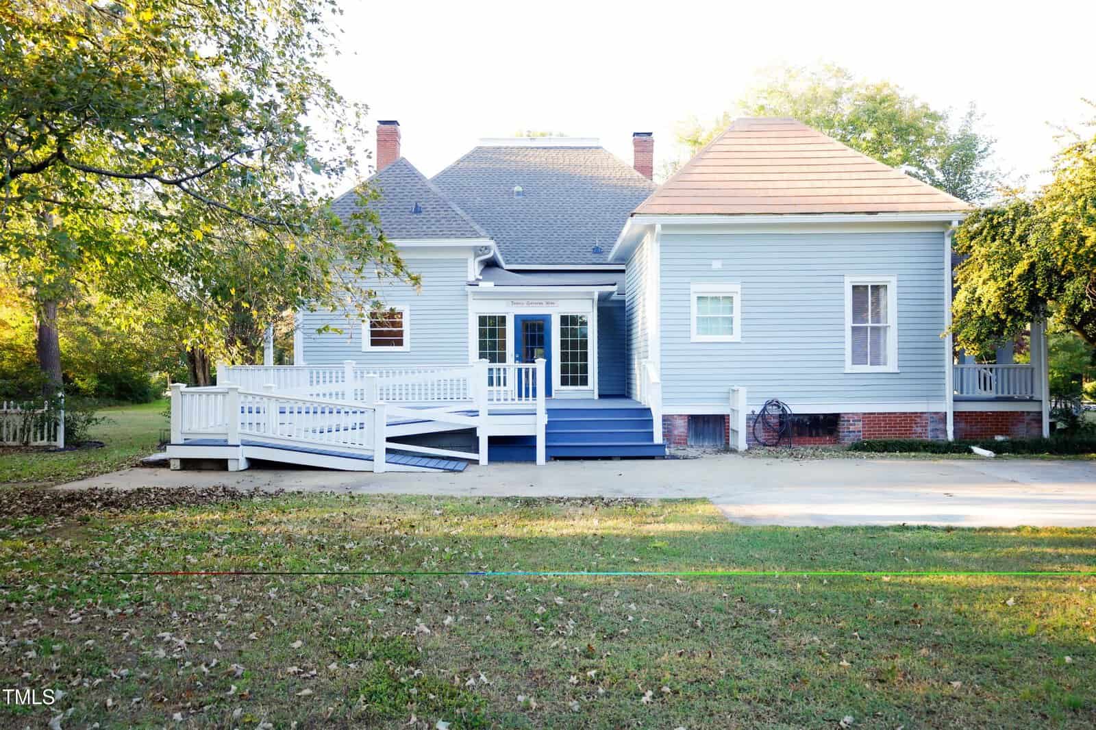 A light blue house with white trim, a ramp leading to a small raised porch, several windows, a brick foundation, and a grassy yard surrounded by trees.