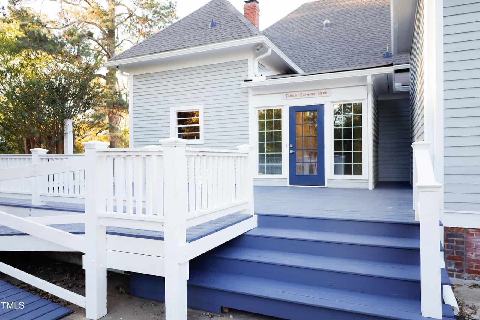 A light gray house with a blue door and matching blue steps leads to a white-railed porch. Large windows are on both sides of the door. Trees and sunlight are visible in the background.