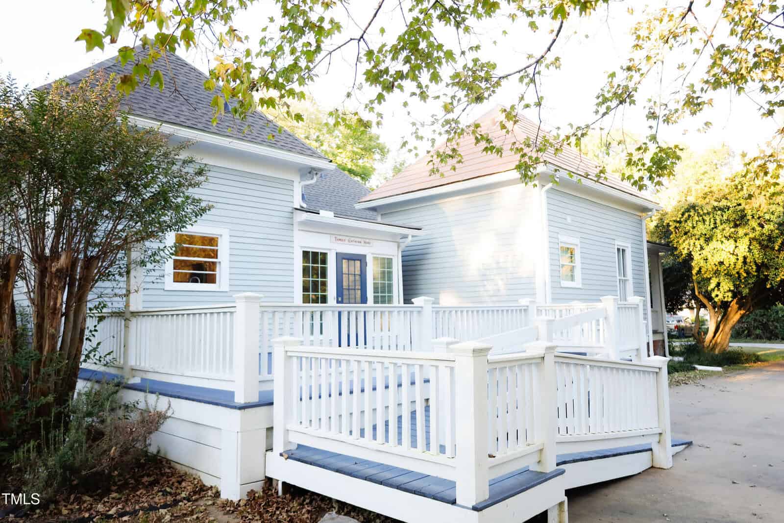 A light blue house with white trim and a front ramp is surrounded by trees with green and yellow leaves. The house has multiple windows and sits on a concrete driveway with some fallen leaves.