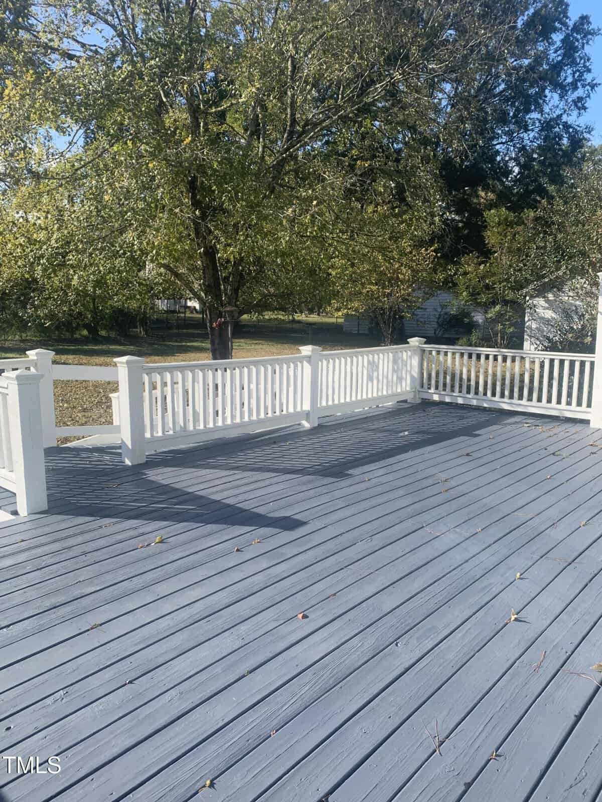 A spacious gray wooden deck with white railings, overlooking a grassy yard with trees in the background. Scattered leaves are visible on the deck surface. The scene is brightly lit by sunlight.