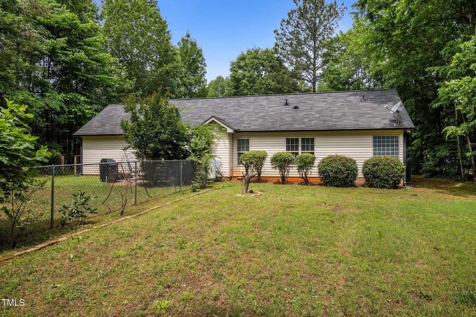 Single-story house with light-colored siding, dark shingles, fenced yard, trimmed bushes along the front, and surrounded by trees on a grassy lawn under a clear blue sky.
