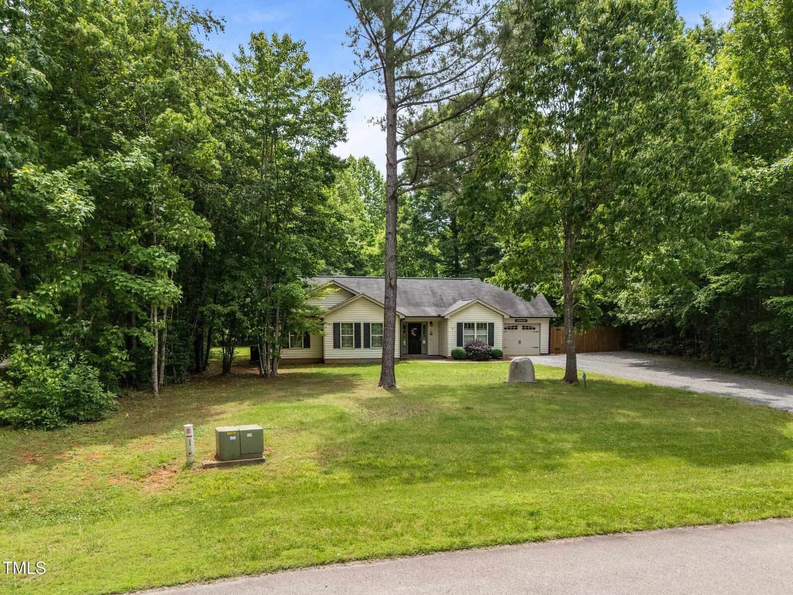 A single-story house with light-colored siding and a dark roof sits surrounded by trees. A long driveway leads to an attached garage, and a large grassy yard extends to the street with a utility box near the curb.