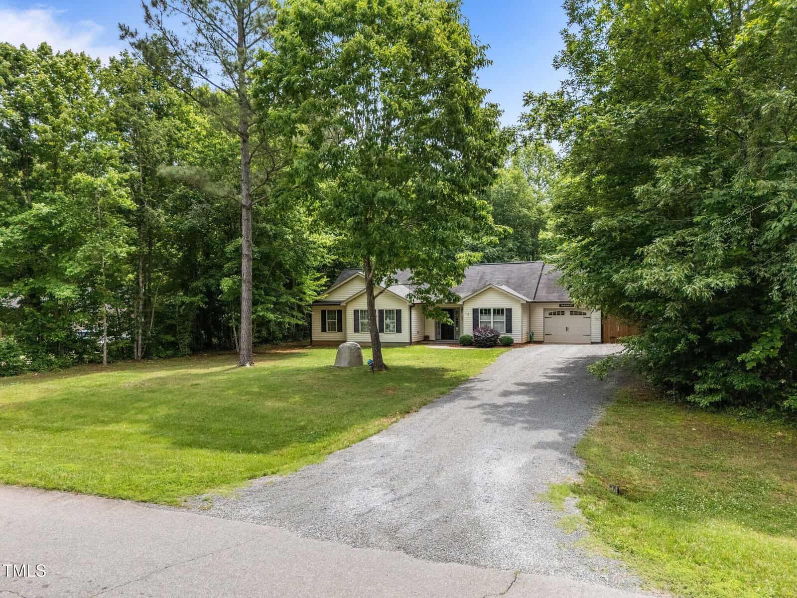 Single-story house with light-colored siding and dark shutters sits surrounded by large trees and greenery. A gravel driveway leads up to the attached garage. The lawn is spacious with a stone marker near the front.