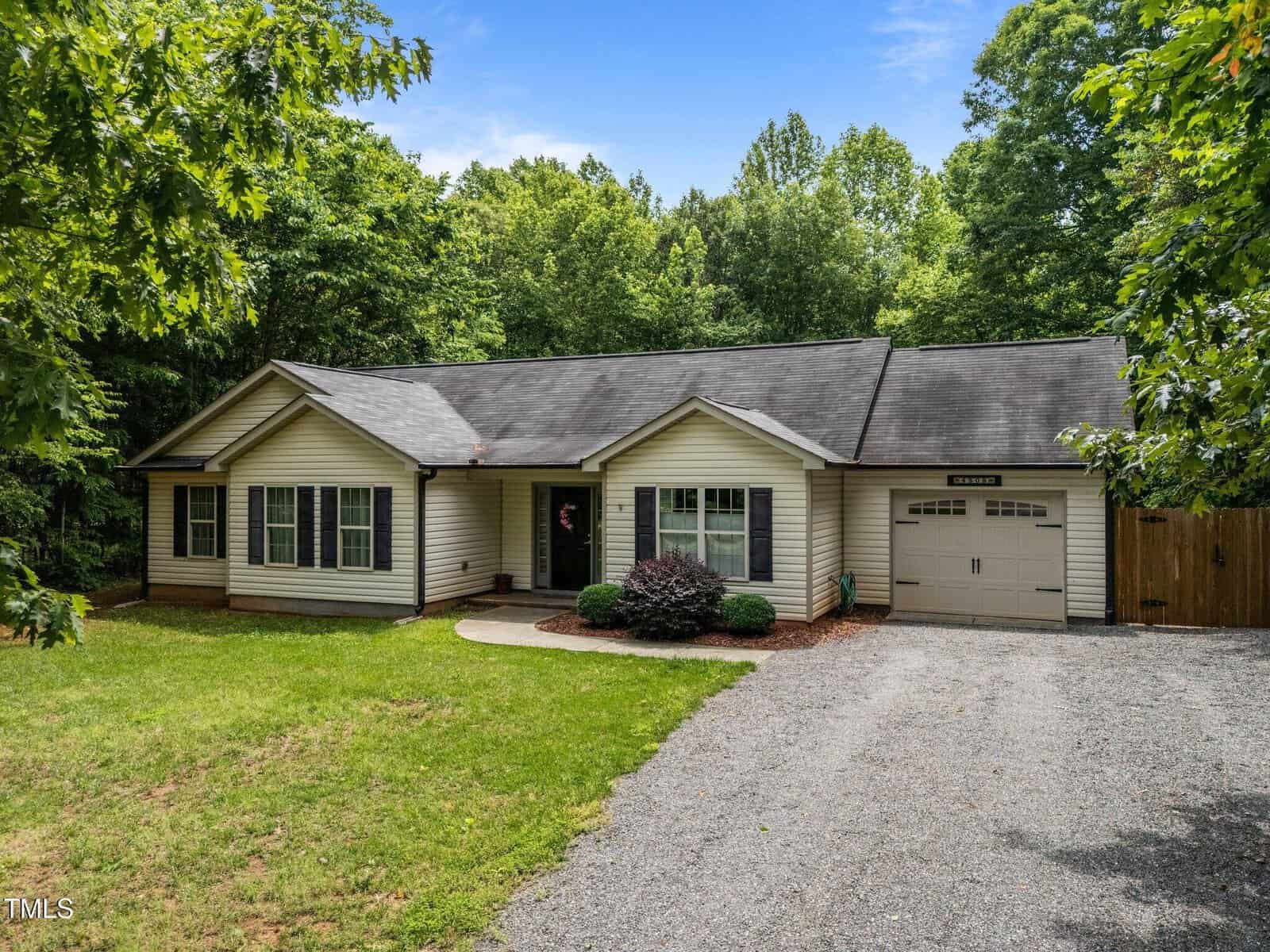 Single-story beige house with black roof, attached one-car garage, and a gravel driveway, surrounded by green trees and a lawn under a blue sky.