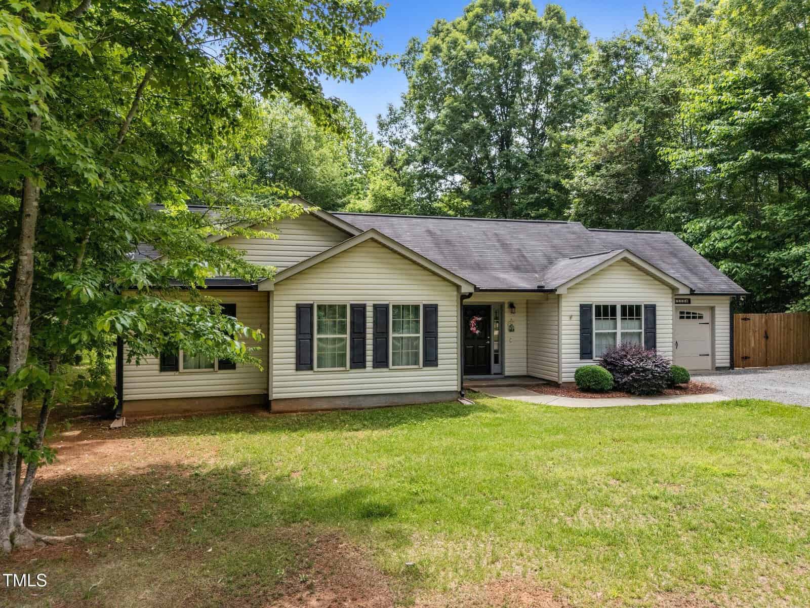 Single-story house with light siding, dark shutters, and a black roof. There are several windows on the front, a single-car garage to the right, and a driveway with a lawn and trees surrounding the property.