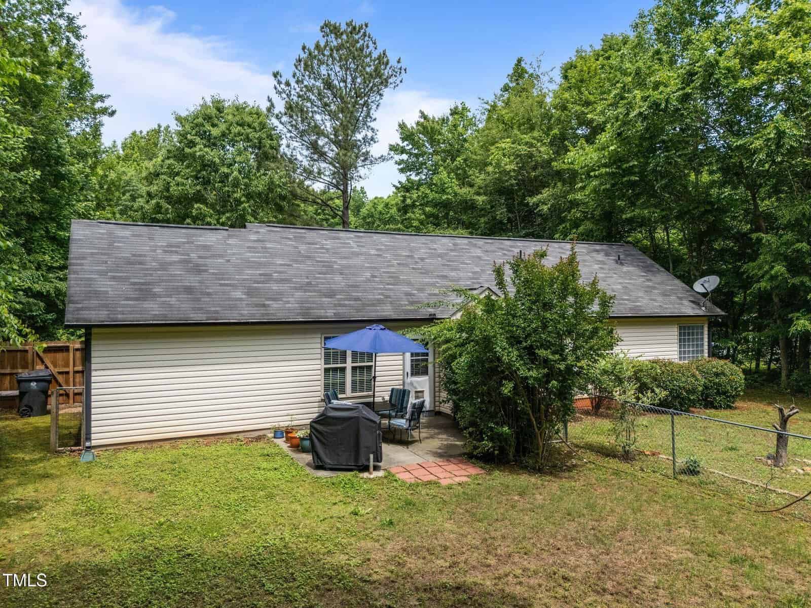 Single-story white house with a dark roof, surrounded by trees and grass. There is a small paved patio with outdoor furniture and a closed blue umbrella. A grill and a chain-link fence are visible on the right.