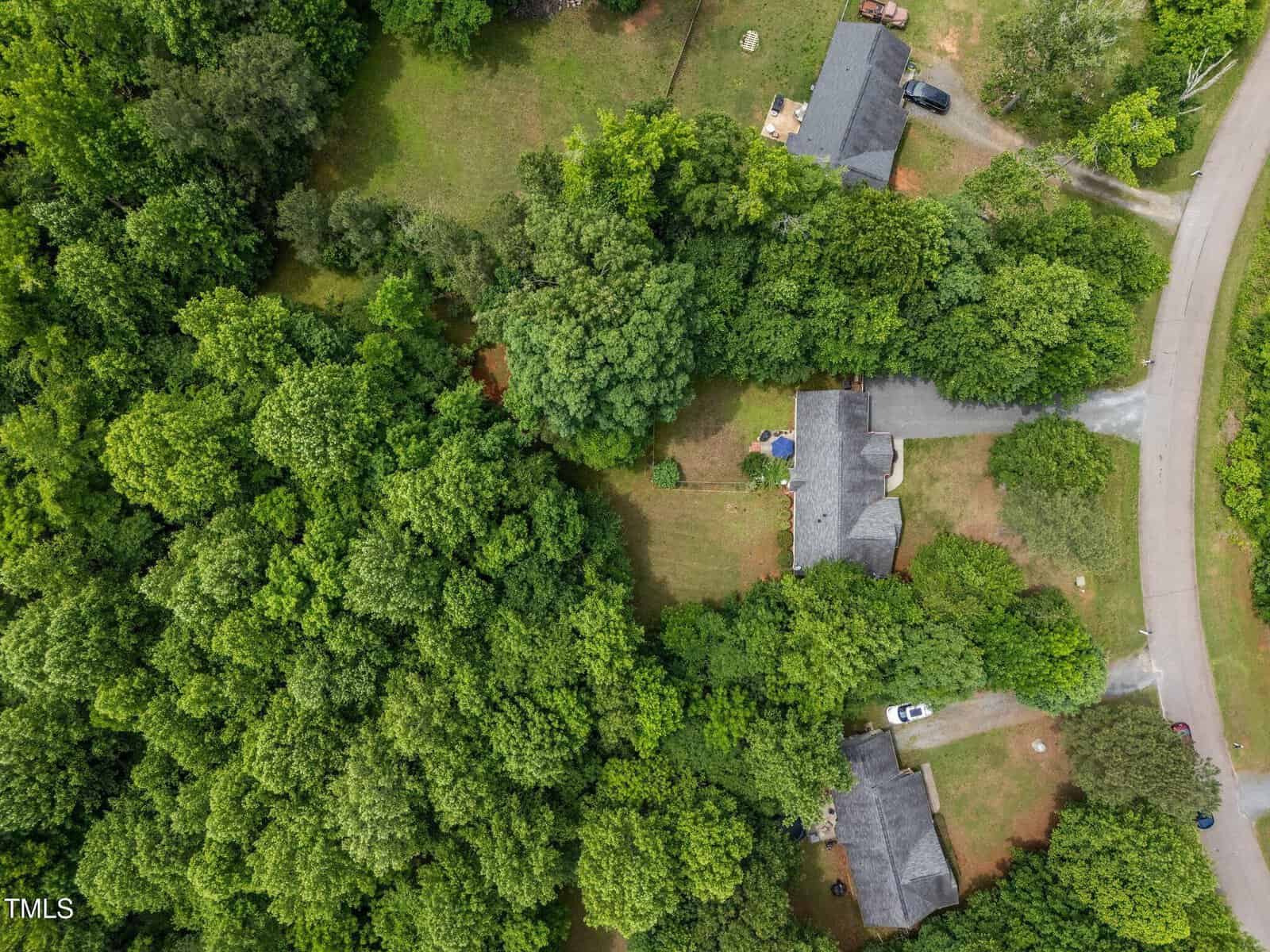 Aerial view of a residential property surrounded by dense green trees, with a lawn area, two driveways, parked vehicles, and a curved road visible on the right side of the image.
