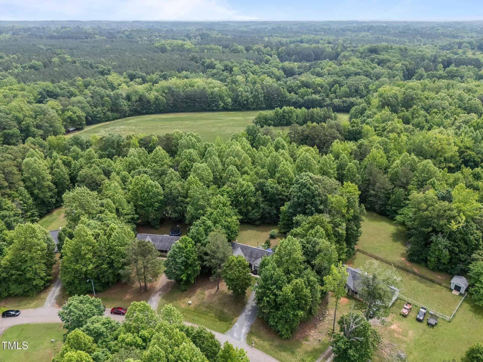 Aerial view of a rural property with several houses surrounded by dense green trees, with open fields and forests in the background.