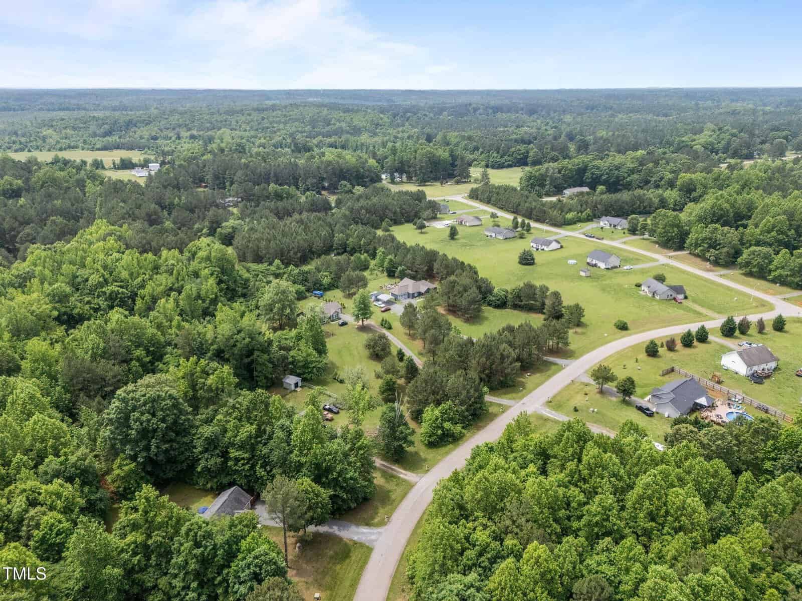 Aerial view of a rural neighborhood with scattered houses, winding roads, dense green trees, and open grassy areas under a clear sky.