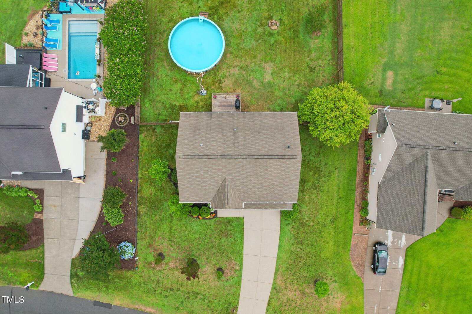 Aerial view of a suburban neighborhood showing three houses, two driveways, a backyard with a round above-ground pool, a rectangular in-ground pool, and green lawns separating each property.