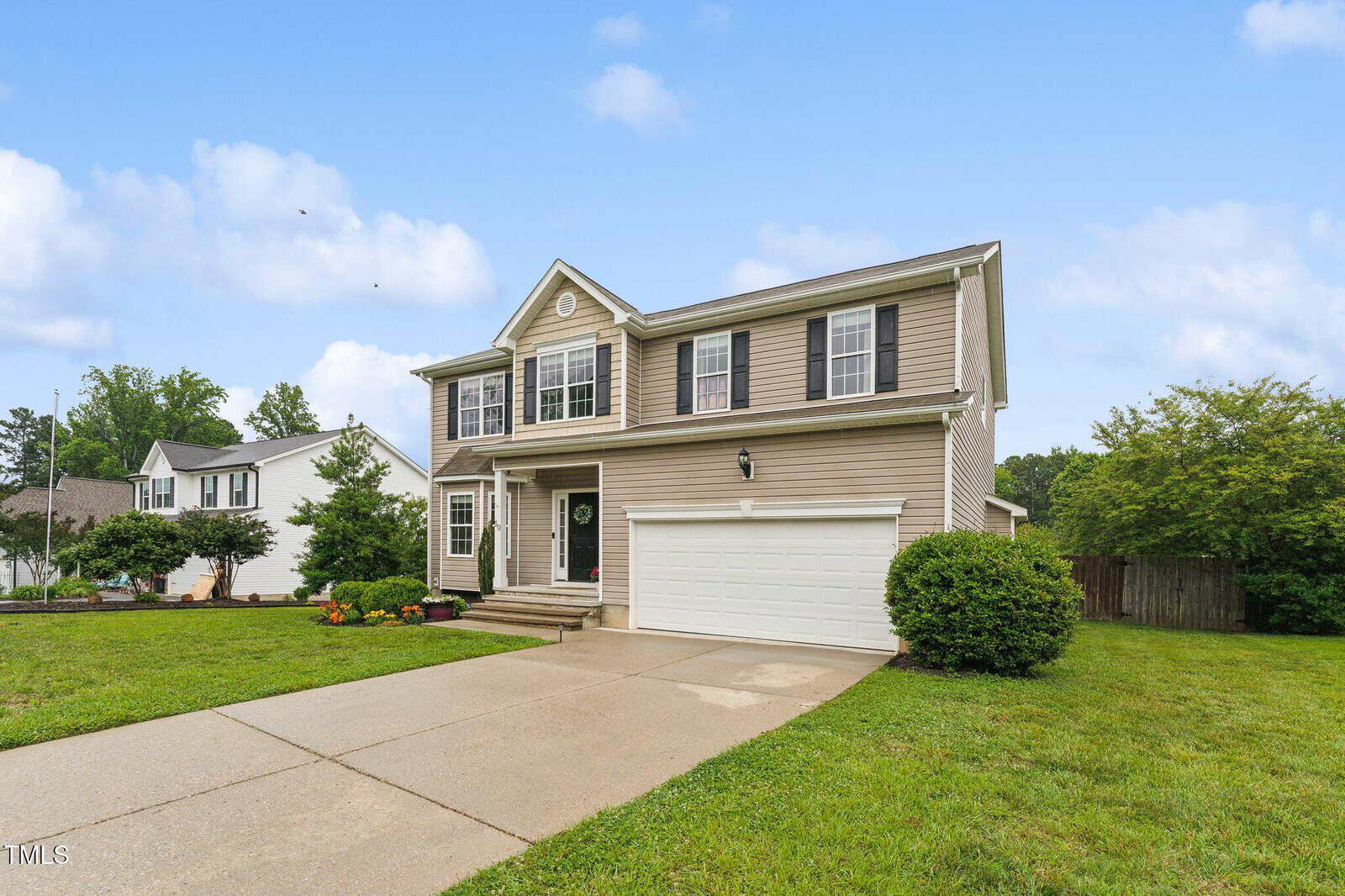 Two-story beige suburban house with white trim and black shutters, a double garage, a concrete driveway, green lawn, small bushes, colorful flowers, and a partly cloudy blue sky in the background.