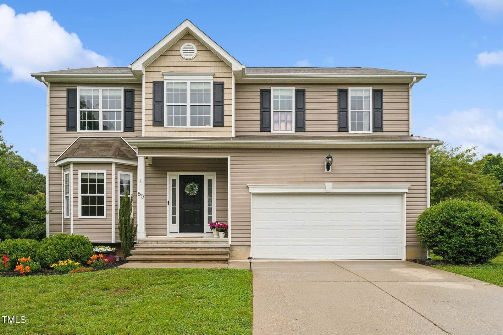 A two-story beige house with white trim and black shutters, featuring a double garage, front porch with potted plants, and a neatly landscaped yard under a blue sky.