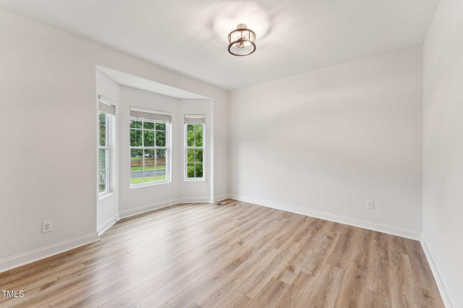 Empty room with light-colored walls, wood-look flooring, a ceiling light fixture, and a bay window letting in natural light. The view outside shows greenery and a lawn. No furniture or decor is present.