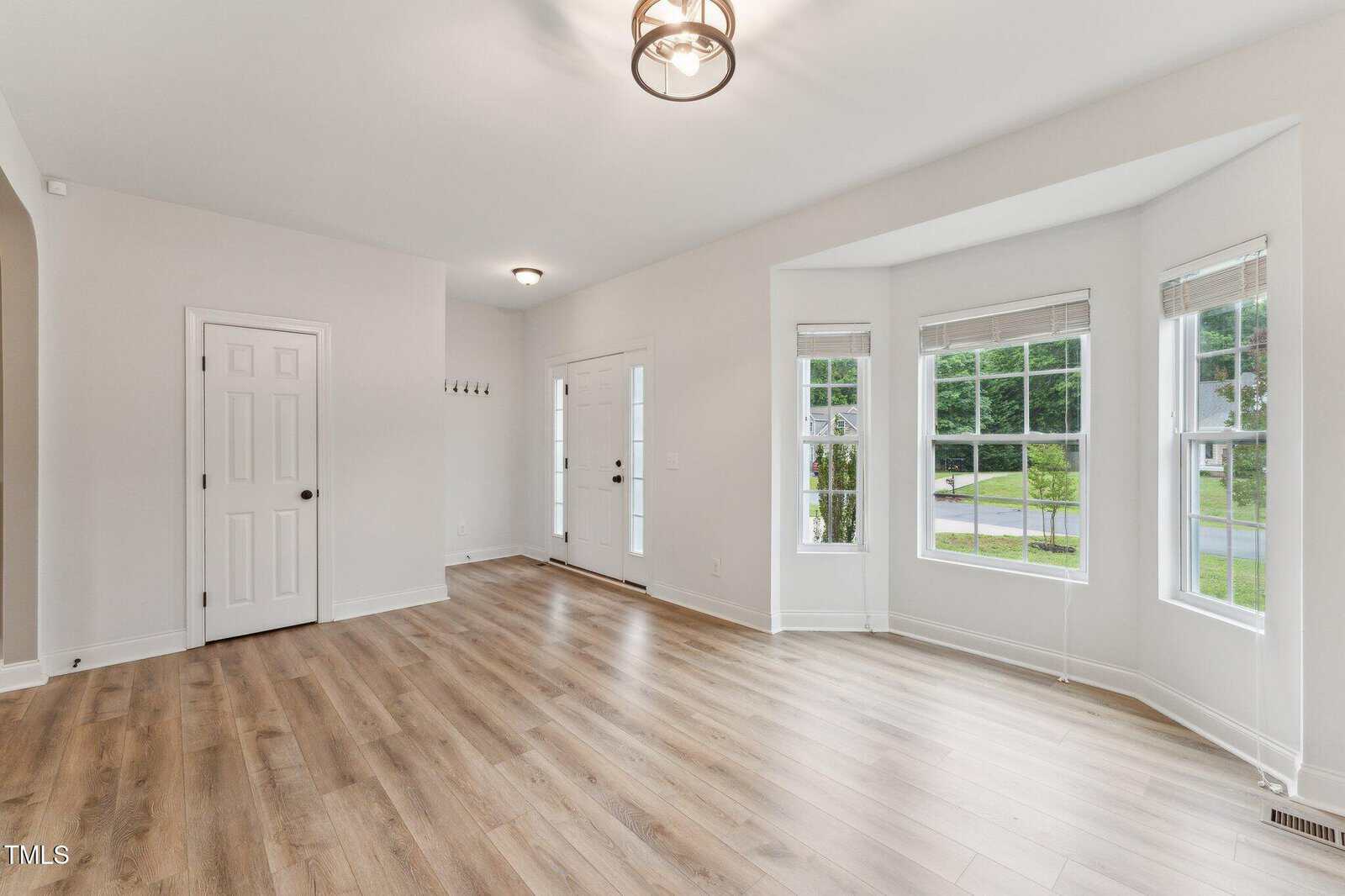 Bright, empty room with light wood floors, white walls, a bay window, two white doors, and a modern ceiling light fixture. Sunlight streams in through the large windows, illuminating the space.