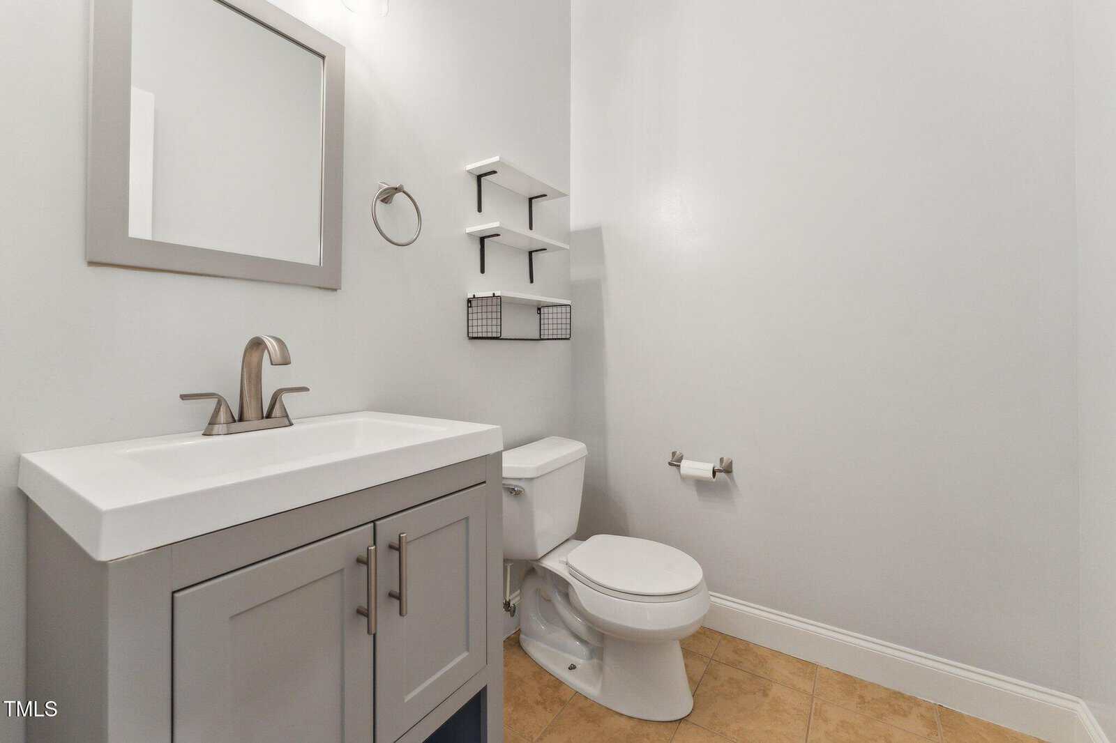 A small bathroom features a gray vanity with a sink, a wall-mounted mirror, a toilet, tile flooring, and white walls. Shelves and a wire basket are mounted above the toilet. A towel ring is next to the sink.