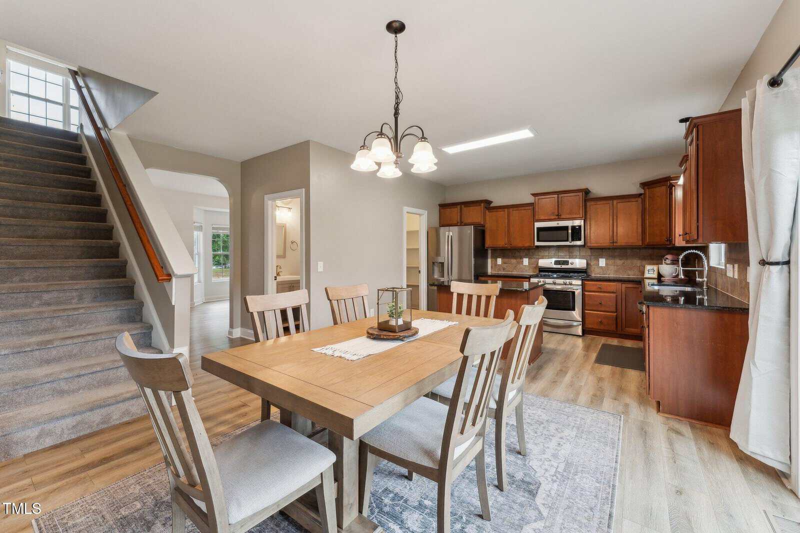 A dining area with a wooden table and six chairs sits next to a kitchen with brown cabinets, stainless steel appliances, and an island. A carpeted staircase is visible on the left. Light wood flooring covers the space.