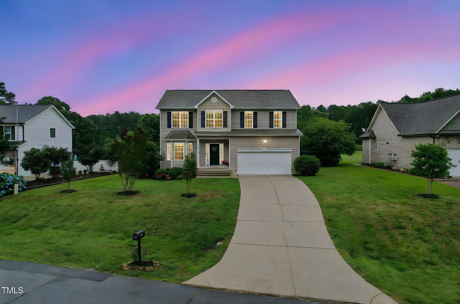 Two-story suburban house with a front porch, illuminated windows, and an attached garage, sits on a green lawn at dusk with a pink and blue sky in the background.