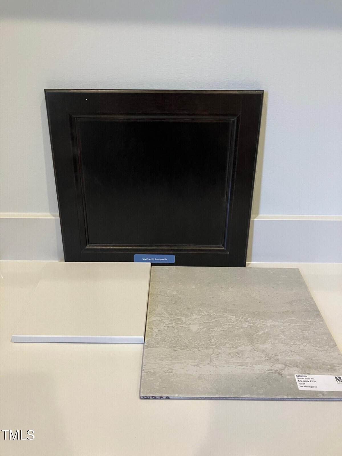 A dark wooden cabinet door sample stands upright behind two square tile samples, one white and one light gray with a textured pattern, displayed on a white countertop against a light-colored wall.