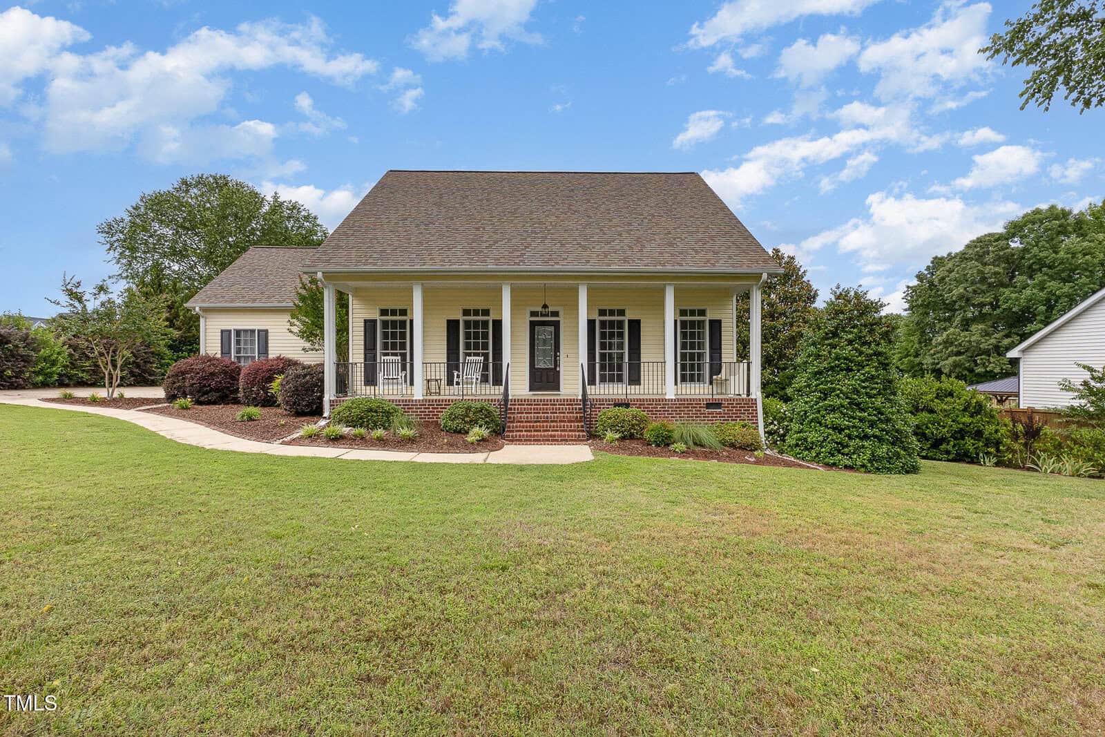 A charming yellow house with a large front porch, black shutters, and white columns, surrounded by neatly trimmed bushes and a spacious green lawn under a blue sky with scattered clouds.
