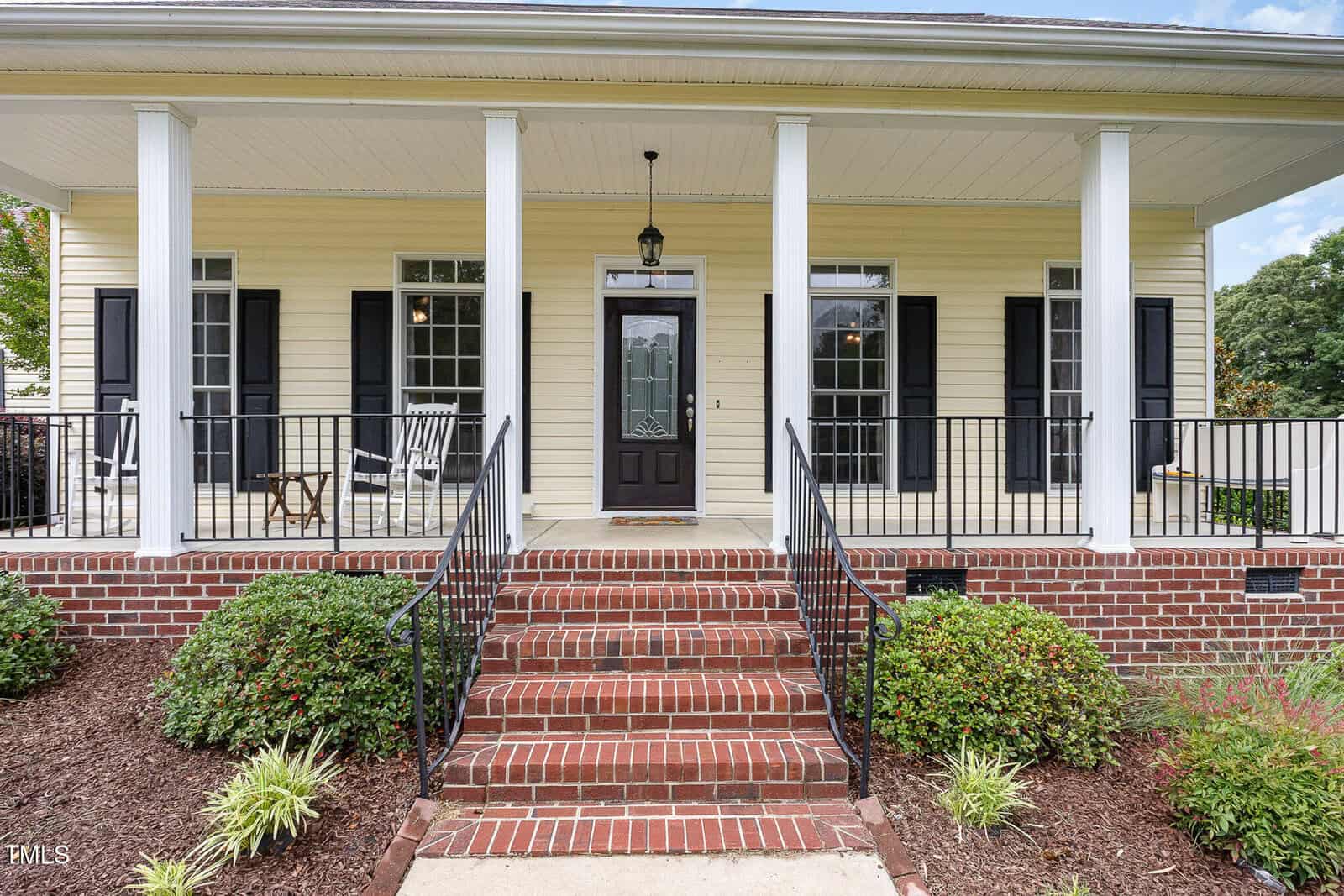 A brick staircase leads to the front porch of a yellow house with white columns, black shutters, and a black front door. Landscaping with shrubs and a white rocking chair are visible on the porch.