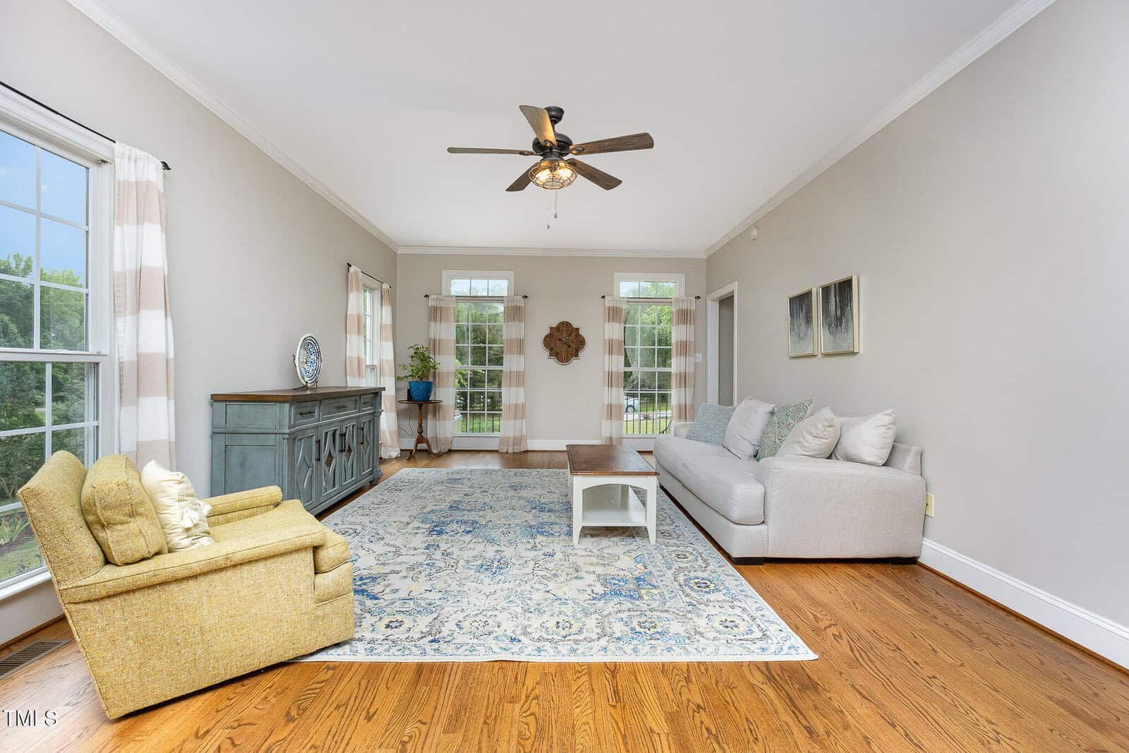 A bright living room with large windows, light gray walls, hardwood floors, a ceiling fan, a gray sofa, a patterned blue rug, a yellow armchair, and a teal sideboard with plants and wall art.