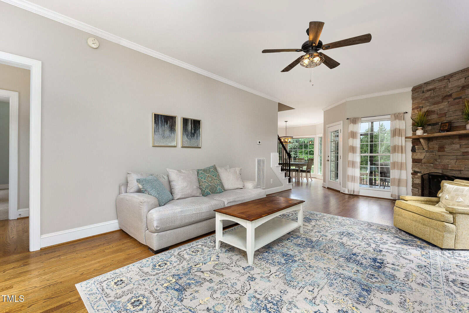A bright living room with a gray sofa, wooden coffee table, light blue patterned rug, and yellow armchair near a stone fireplace. Ceiling fan and large windows with glass doors let in natural light.