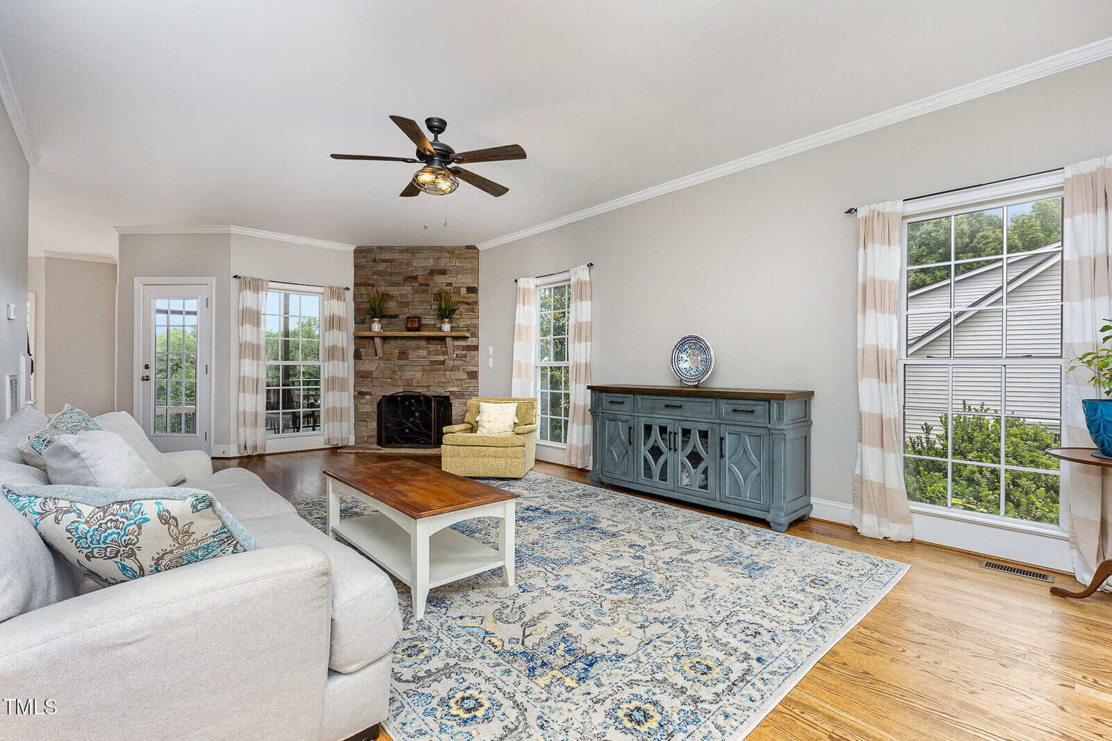 A living room with a white sofa, wooden coffee table, blue patterned rug, fireplace with brick surround, ceiling fan, blue sideboard, large windows with curtains, hardwood floor, and a yellow armchair.