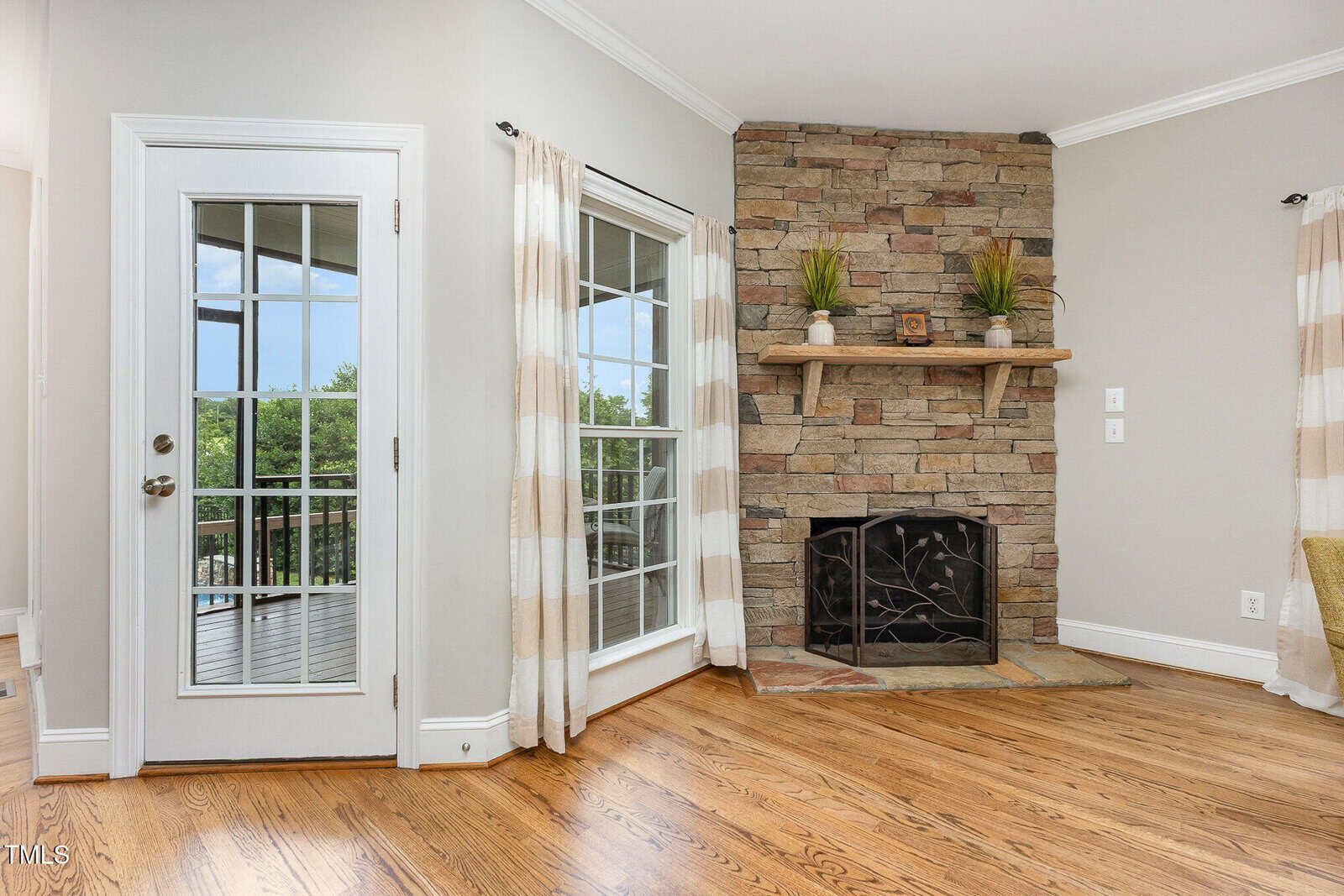 A room with a wooden floor, stone fireplace with a mantle, glass door, and large windows with beige-and-white curtains. The fireplace has a metal screen and is decorated with plants and small items.