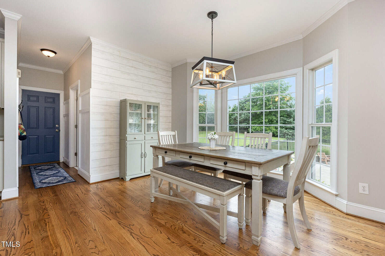 Bright dining area with a wooden table, chairs, and bench, placed by large corner windows. Light gray walls, white trim, hardwood floor, and a modern geometric light fixture overhead. Entryway with blue door visible.