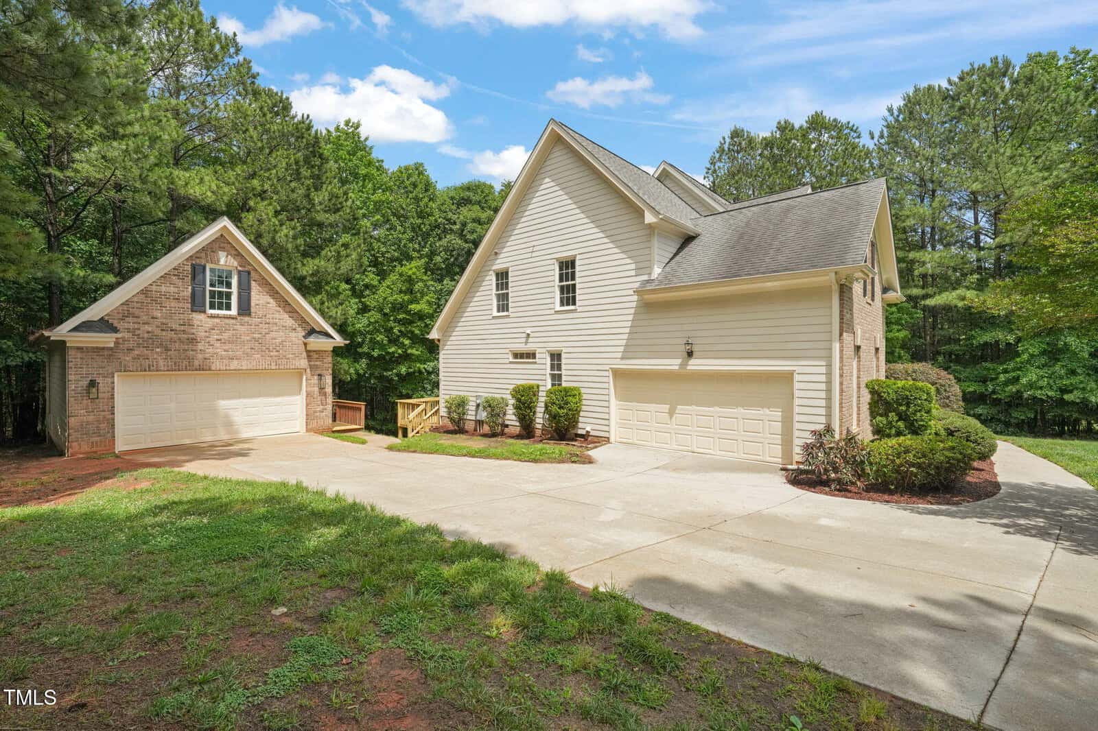 A two-story house with beige siding and a three-car garage, next to a separate brick two-car garage, surrounded by trees. A concrete driveway connects both garages. The sky is partly cloudy.
